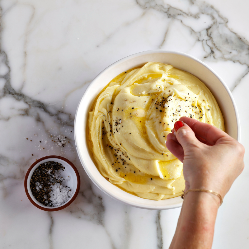 Hand sprinkling black pepper over mashed potatoes in a white bowl on a marble surface