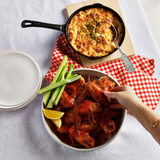 Bowl of buffalo chicken wings with a side of baked dip on a checkered tablecloth.