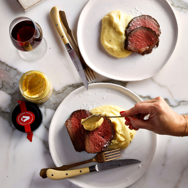 Two plates of sliced beef with mashed potatoes, a glass of red wine, and a jar of mustard on a marble surface.