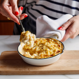 Person serving macaroni and cheese from a bowl on a wooden cutting board.