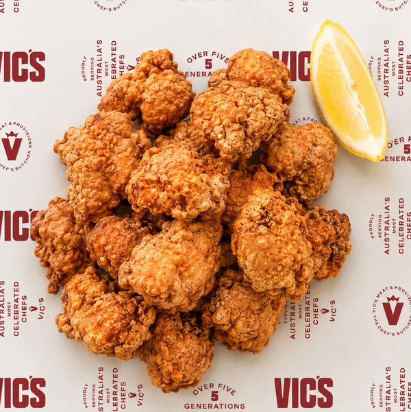 A pile of golden-brown fried chicken pieces on branded paper, with a lemon wedge on the right side. The paper has a repeating Vic's logo pattern.
